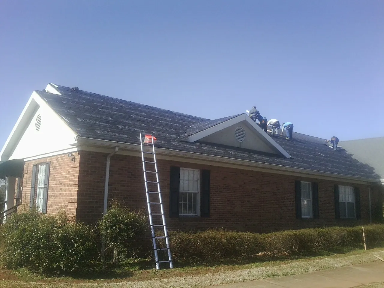 Crew tearing shingles off a brick ranch roof with a ladder set — Metal Roof Repair work by our crew in Swanton