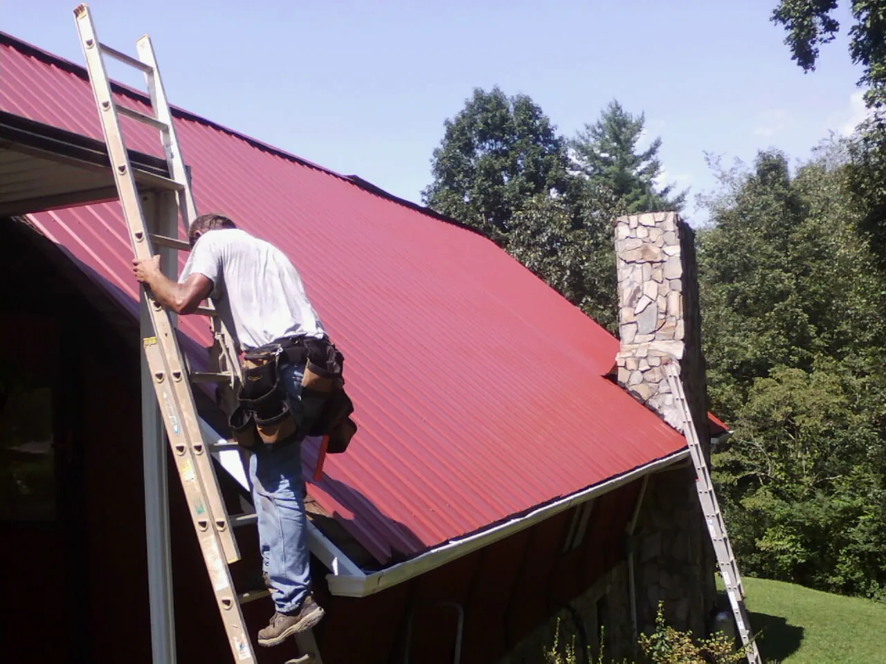 Roofer on a ladder beside a red metal roof and stone chimney — Soffit & Fascia Repair by Roofing Brothers in Swanton